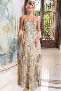 A woman models a strapless floor-length gown with a beige and silver floral pattern, sheer draped sleeves, and a matching sheer headpiece in an elegant indoor setting.