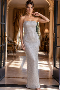 A woman models a strapless, floor-length silver gown with a textured, beaded surface in an elegant, sunlit room with arched doorways and warm drapery.