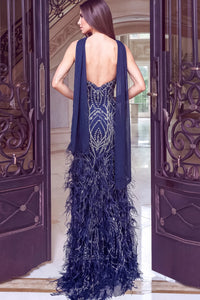 A woman stands in a grand doorway wearing a navy blue evening gown with a sheer draped overlay and feathered skirt details, shown from the back.