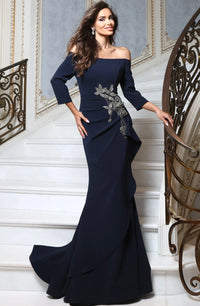 A woman models an off-the-shoulder navy blue evening gown with three-quarter sleeves and a silver-gray floral applique on the bodice, standing on white stairs in an elegant interior.