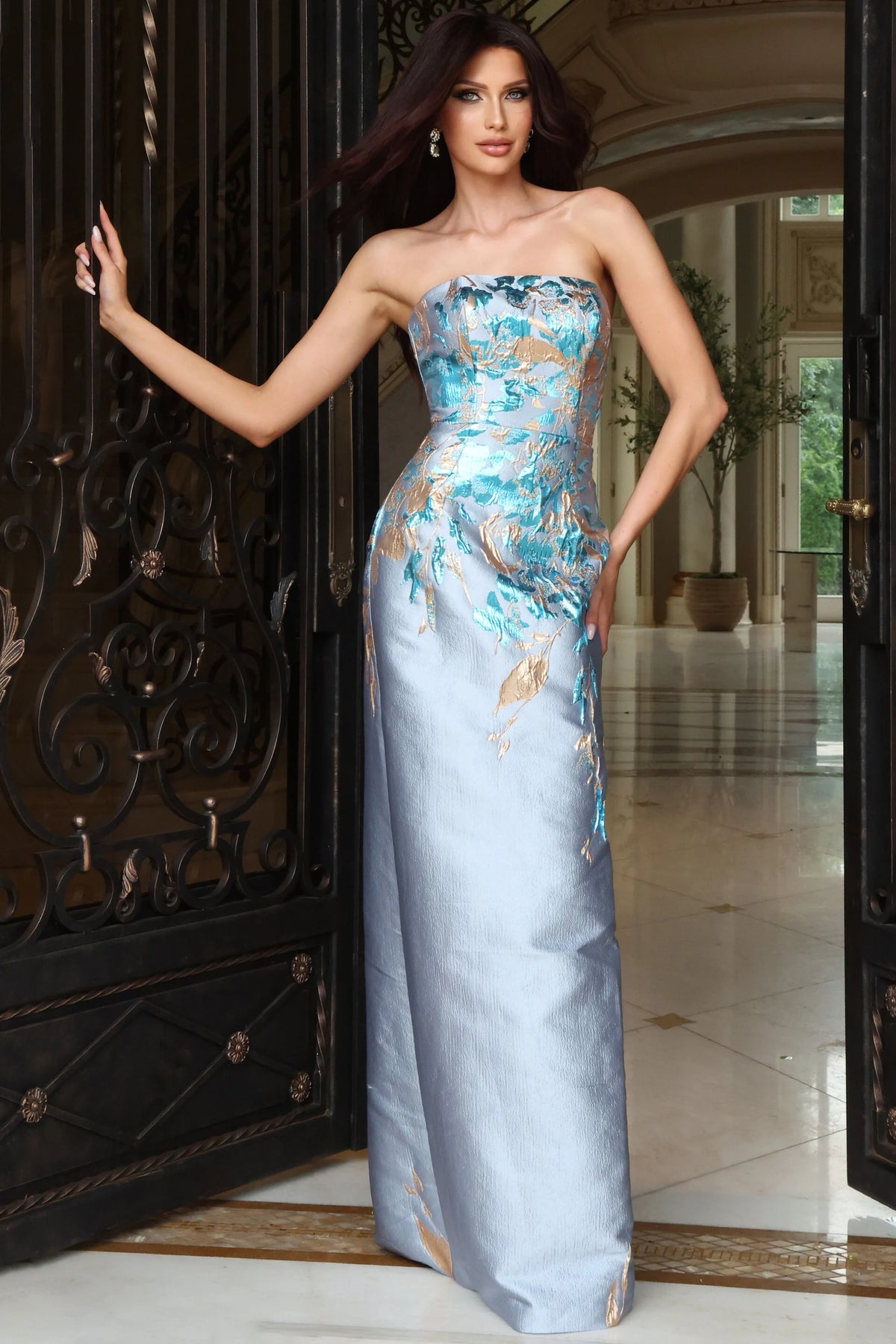 A woman models a strapless formal gown in a grand doorway. The gown features a light blue satin skirt with gold and teal abstract floral patterns on the bodice and cascading down the sides.