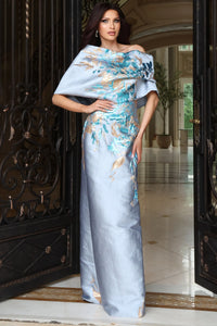 A woman models a floor-length off-the-shoulder gown in light blue satin with elaborate gold and teal floral embroidery, standing in an elegant hallway framed by open black metal doors.