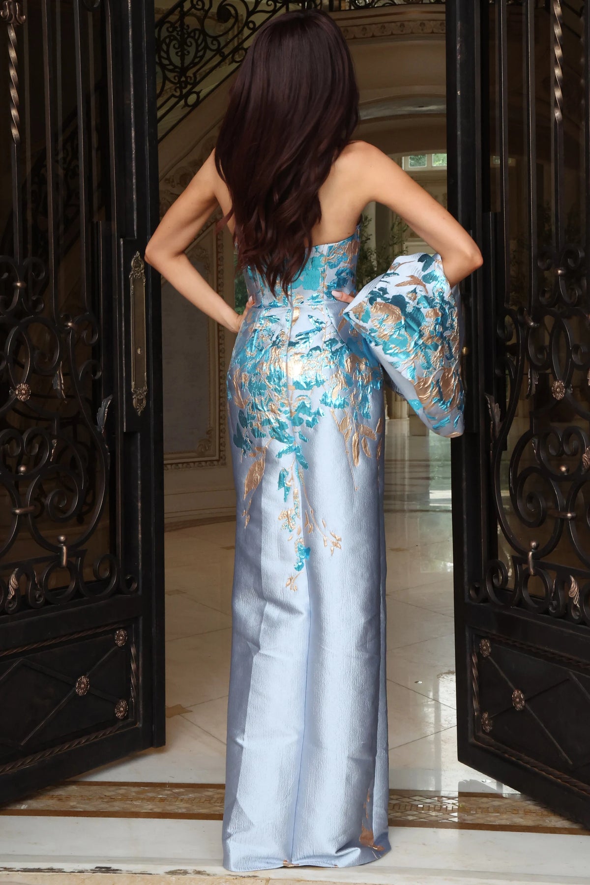 A woman stands with her back to the camera wearing a strapless light blue satin gown with a large floral print in turquoise, gold, and white. She is posed in front of open dark wrought-iron doors.