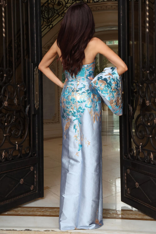 A woman stands with her back to the camera wearing a strapless light blue satin gown with a large floral print in turquoise, gold, and white. She is posed in front of open dark wrought-iron doors.