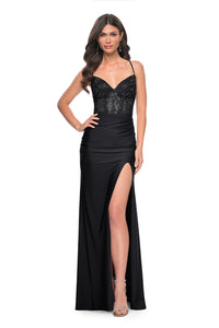 La Femme 31988 Rhinestone Embellished Lace Bodice Prom Gown - A glamorous long dress featuring a ruched jersey skirt, high slit, rhinestone embellished lace bodice with exposed boning, lace-up back, and ruching along the zipper for a perfect fit. The model is wearing the dress in black.