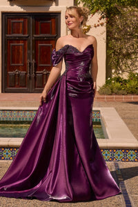 A woman models a floor-length off-the-shoulder formal gown in rich eggplant color, standing outdoors in front of a building with a tiled pool and wooden doors.