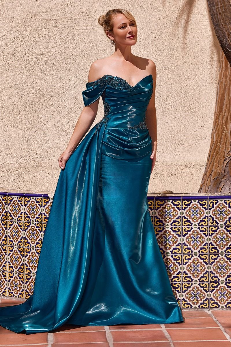 A woman models a floor-length peacock color gown with an off-the-shoulder neckline, lace bodice, ruched waist, and a full skirt, standing against a beige stucco wall with patterned tile and a tree trunk in the background.