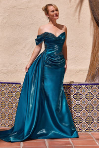 A woman models a floor-length peacock color gown with an off-the-shoulder neckline, lace bodice, ruched waist, and a full skirt, standing against a beige stucco wall with patterned tile and a tree trunk in the background.