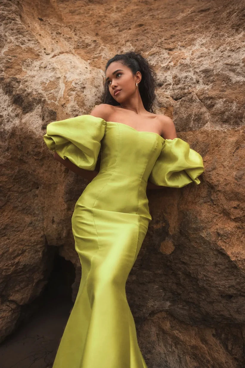 Model wearing Mac Duggal 50677 off-the-shoulder satin gown with puff sleeves, mermaid silhouette, and sweeping train.  Model is outdoors wearing the color chartreuse. Front view.
