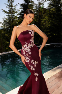 Model wearing Nicoletta NC2080 strapless black gown with blush floral embroidery in a mermaid silhouette by a pool.  Model is wearing the dress in the color burgundy.