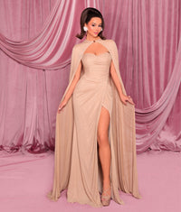Model wearing Nicoletta NC3004 gown featuring a draped cape, strapless bodice, and front slit, posed against a pink curtain backdrop.