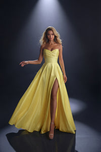 A woman modeling a strapless lemon yellow formal gown with a sweetheart neckline, fitted bodice, full skirt, and high front slit, photographed on a dark studio background with a spotlight.