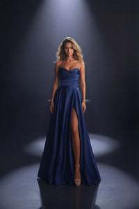 A woman stands in a dramatic studio setting wearing a strapless navy blue satin gown with a sweetheart neckline, ruched bodice, high thigh slit, and floor-length skirt under spot lighting.