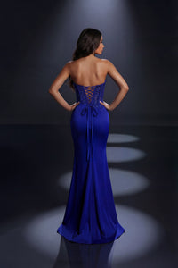 A woman stands with her back to the camera wearing a strapless cobalt blue evening gown with lace-up back detailing, photographed in a dark studio with spotlight effects.