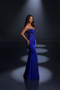 A woman modeling a strapless cobalt blue evening gown with a beaded corset-style bodice and fitted silhouette, photographed in a dark studio with spotlights on the floor.