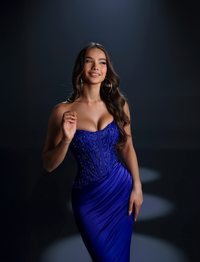 A woman modeling a strapless cobalt blue evening gown with a heavily embellished corset-style bodice and draped skirt, photographed against a dark studio background with spotlights.