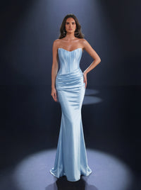 A woman modeling a strapless light blue satin evening gown with a structured corset-style bodice and a fitted mermaid silhouette, photographed in a dark studio with spotlights.