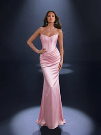 A woman models a strapless pink satin evening gown with a structured corset-style bodice and ruched hips, standing against a dark studio background with spotlights.