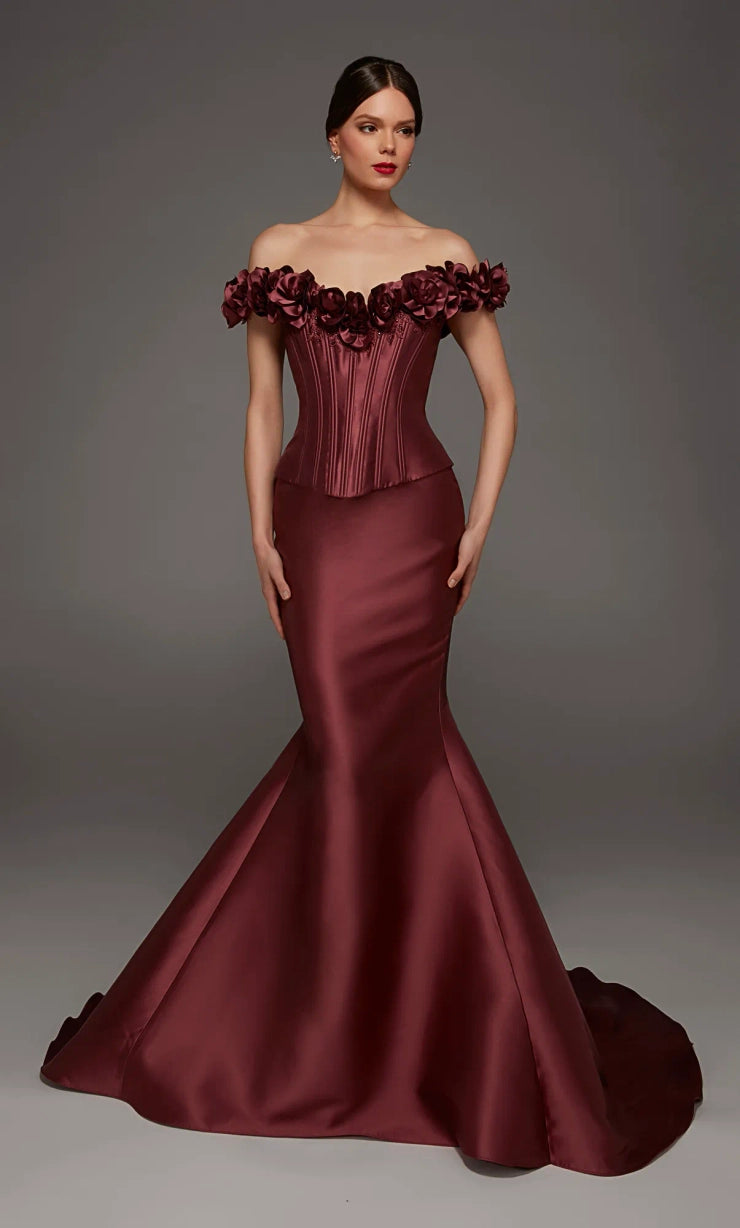 A woman modeling an off-the-shoulder mermaid gown in deep burgundy satin, featuring a structured corset-style bodice and a full flared skirt with a train, against a dark gray studio background.