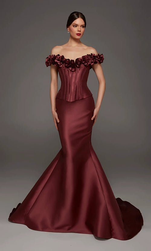 A woman modeling an off-the-shoulder mermaid gown in deep burgundy satin, featuring a structured corset-style bodice and a full flared skirt with a train, against a dark gray studio background.