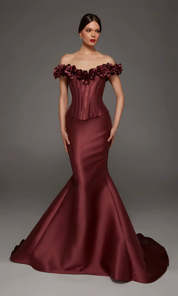A woman modeling an off-the-shoulder mermaid gown in deep burgundy satin, featuring a structured corset-style bodice and a full flared skirt with a train, against a dark gray studio background.