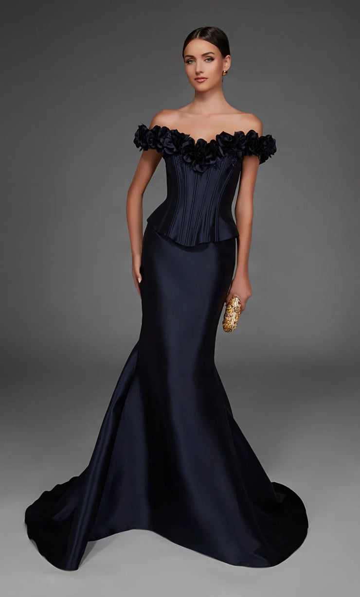 A woman models an off-the-shoulder navy blue evening gown with a structured ruffled bodice, fitted satin skirt, and a slight train, holding a small gold clutch against a dark gray studio background.