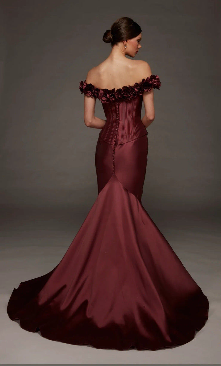 A woman modeling an off-the-shoulder mermaid gown in deep burgundy satin, photographed from the back against a dark gray studio backdrop.