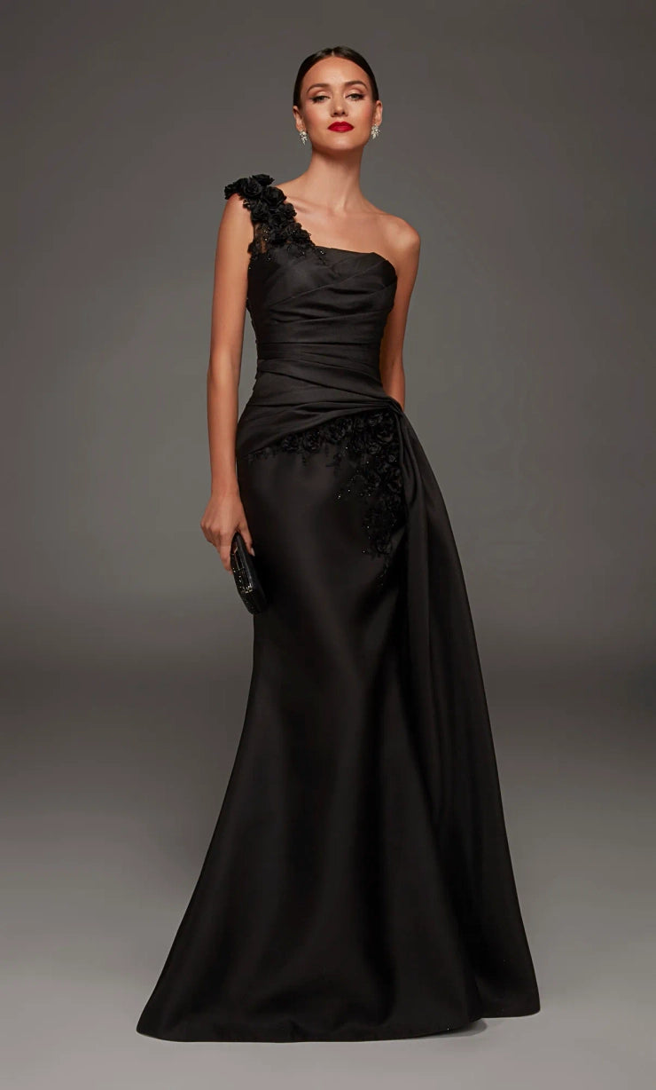 A woman modeling a floor-length black one-shoulder evening gown with a draped satin skirt and floral lace applique details, posed against a dark gray studio background.