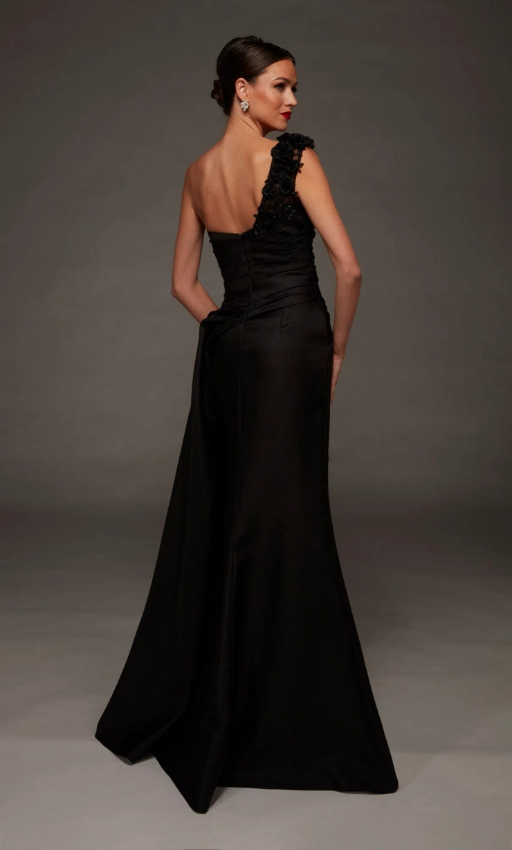 A woman modeling a floor-length black evening gown from the back, featuring a one-shoulder design with ruffled detailing on the shoulder and a structured back, set against a dark gray studio background.