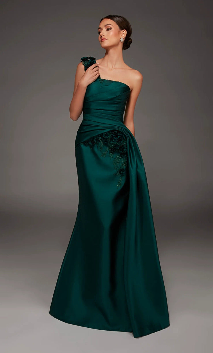 A woman modeling a floor-length, one-shoulder formal gown in deep emerald green satin with a draped bodice and lace applique on the skirt, photographed against a dark gray studio background.