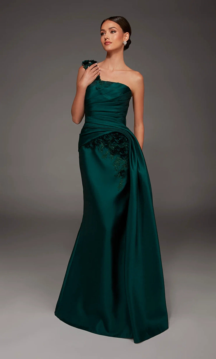 A woman modeling a floor-length, one-shoulder formal gown in deep emerald green satin with a gathered ruched bodice and lace applique at the hip, photographed against a dark gray studio background.