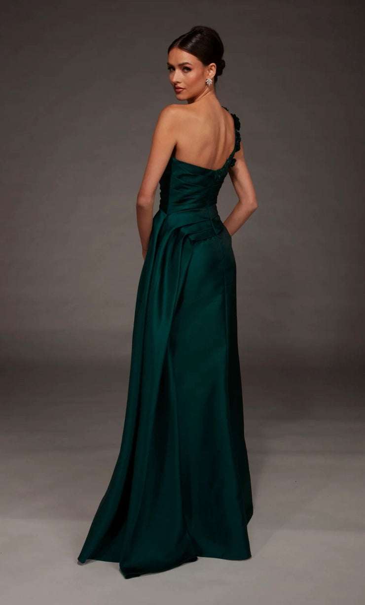 A woman modeling a floor-length dark green satin gown from the back, featuring a one-shoulder strap design and a draped waist, set against a dark gray studio background.