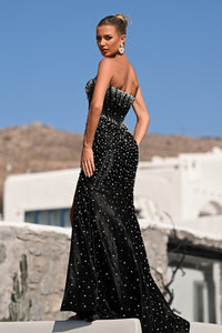 Woman in a black strapless gown with sequins standing against a clear blue sky.