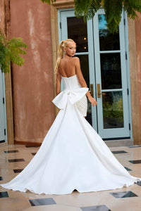 A woman wearing a strapless white wedding gown with a large bow at the back, standing in front of a building with glass doors and tiled flooring.