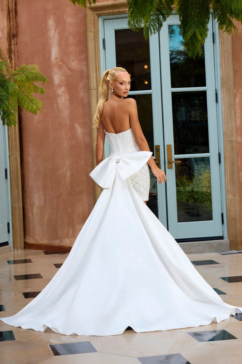 A woman wearing a strapless white wedding gown with a large bow at the back, standing in front of a building with glass doors and tiled flooring.