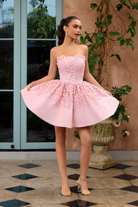Woman in a pink strapless dress standing outdoors with plants and a building in the background.