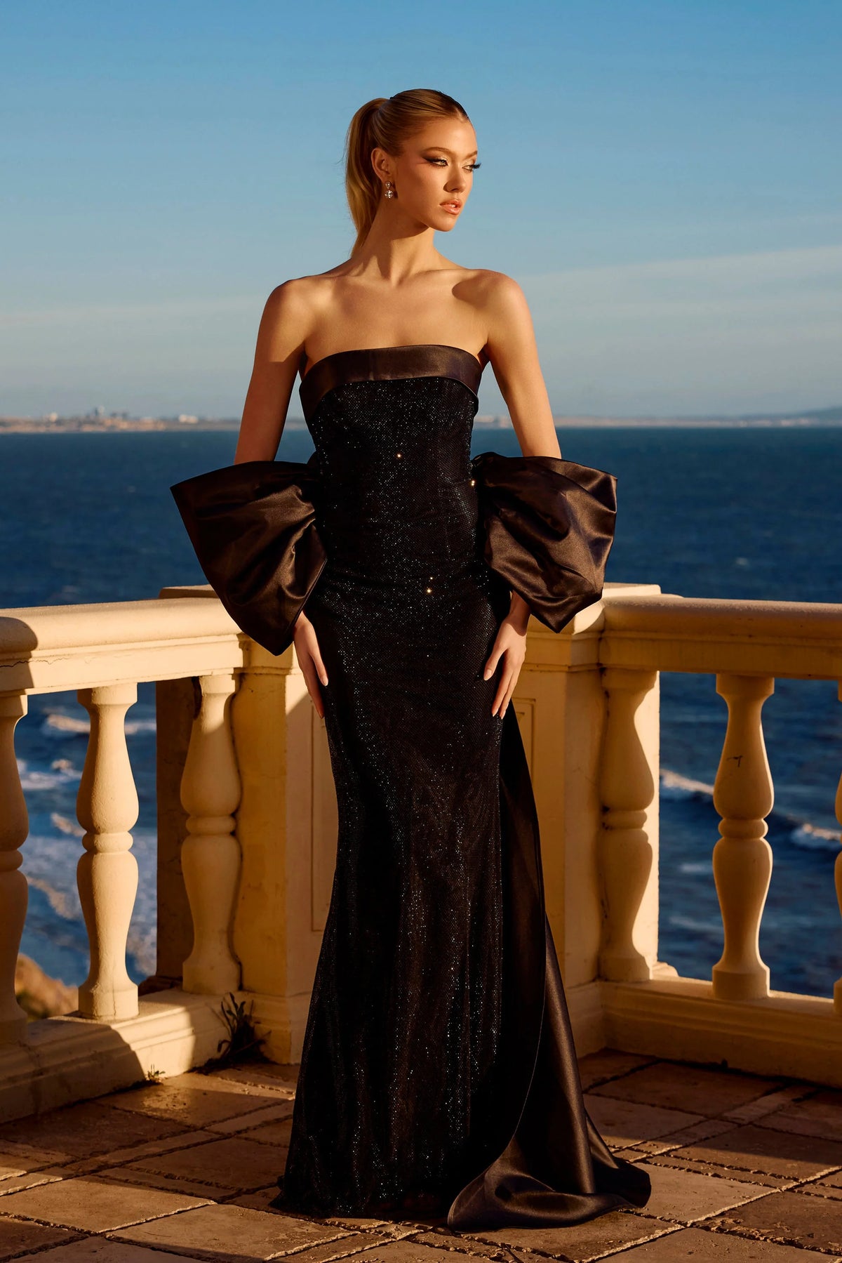 Woman in a black strapless gown with puffed sleeves standing on a balcony overlooking the ocean.