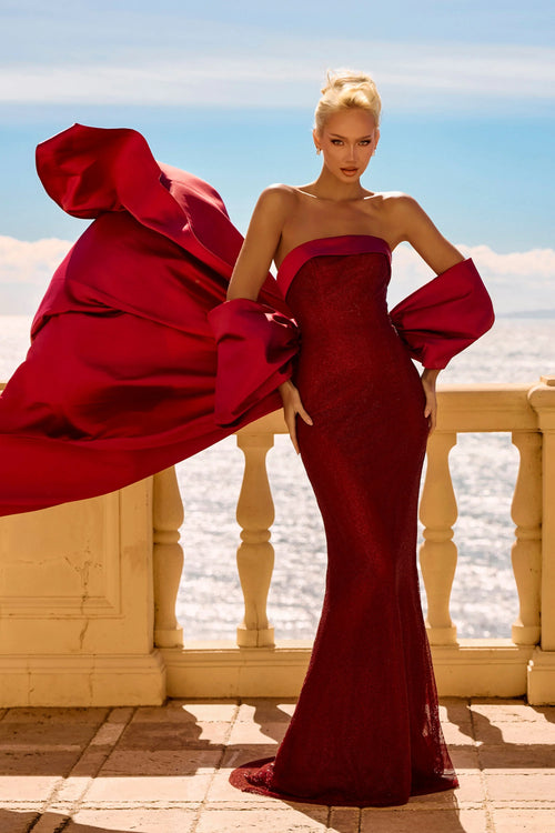 A model stands on a sunlit terrace wearing a strapless deep red evening gown with dramatic oversized sleeves and a flowing train, with a sea and sky background.