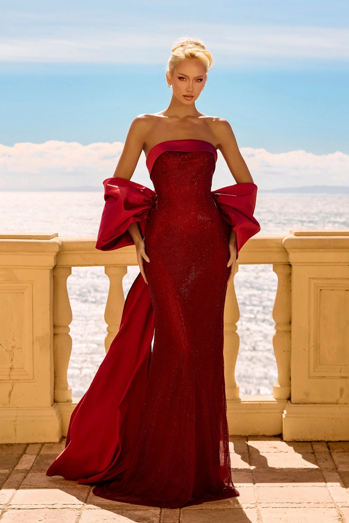 Woman in a red strapless gown with large bow detail standing on a balcony with a scenic background.