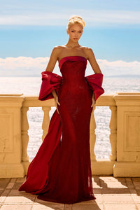 Woman in a red strapless gown with large bow detail standing on a balcony with a scenic background.