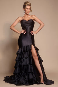 Woman wearing a black strapless gown with ruffled layers and a high slit on a beige background