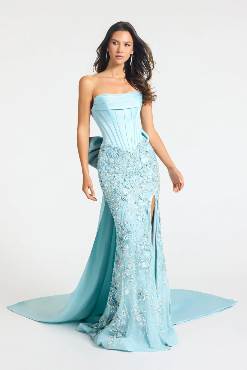 A woman modeling a strapless light blue evening gown with a fitted corset-style bodice, heavily embroidered floral detailing, a thigh-high slit, and a long flowing train against a plain light gray studio background.