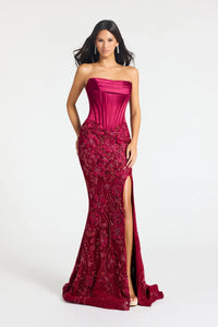 A woman modeling a strapless floor-length gown in rich burgundy. The bodice is smooth and structured, while the skirt features dense floral lace applique and a high front slit with a train, set against a plain light gray studio background.