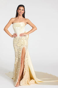 A woman modeling a strapless formal gown with a structured corset-style bodice, fitted lace skirt, high front slit, and a long flowing train against a light gray studio background.