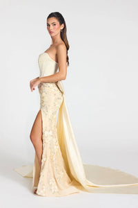 A woman models a strapless cream-colored evening gown with a high thigh slit, floral lace applique, and a long flowing train against a plain light gray studio background.