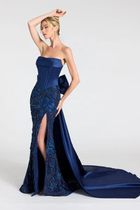 A woman models a strapless floor-length gown in navy blue satin and embellished lace, featuring a high thigh slit and a long draped train against a light gray studio background.