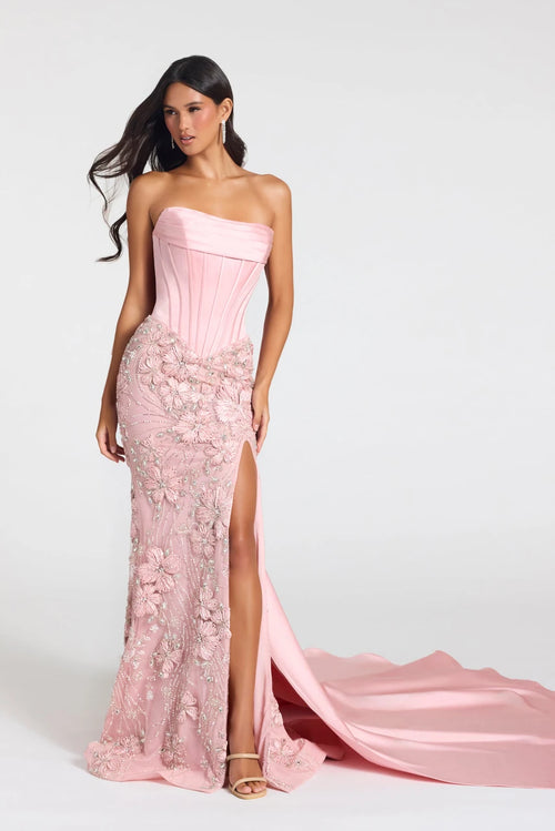 A woman models a strapless pink gown with a structured corset-style bodice, floral lace applique, a high front slit, and a long flowing train against a light gray studio background.