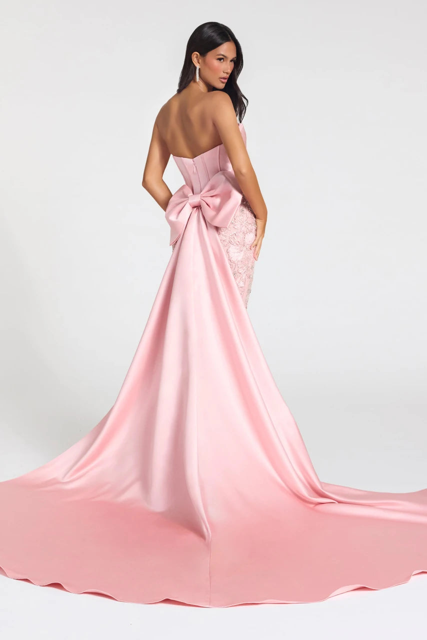 A woman modeling a strapless pink evening gown from the back, featuring a large bow at the back of the bodice and a long satin train on a light gray studio background.