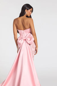 A woman modeling a strapless pink gown from the back, featuring a large satin bow at the waist and lace detailing on the sides, against a light gray studio background.