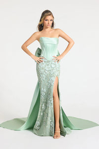 A woman modeling a strapless sage formal gown with a structured corset bodice, floral lace skirt, high thigh slit, and a long draped train against a light gray studio background.